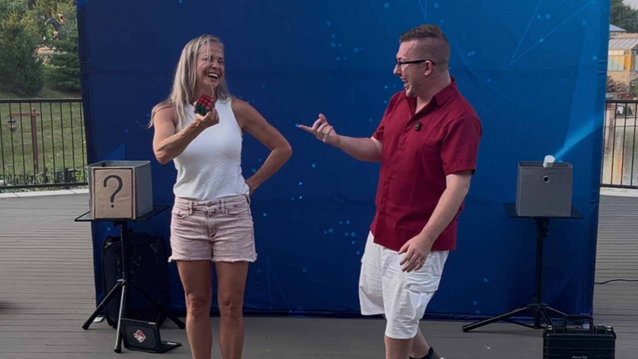 A guest laughs in disbelief holding a solved Rubik's cube while Tyler reacts beside her at an outdoor show