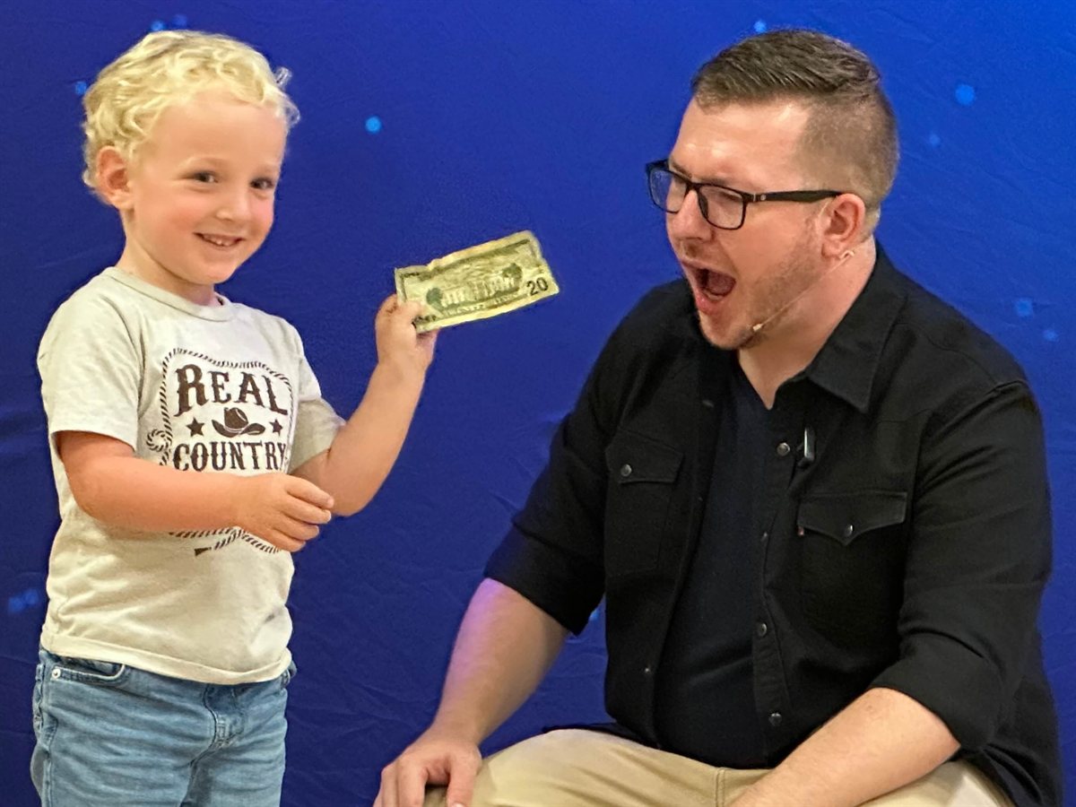 A young volunteer holds up the restored twenty dollar bill while Tyler reacts in mock disbelief