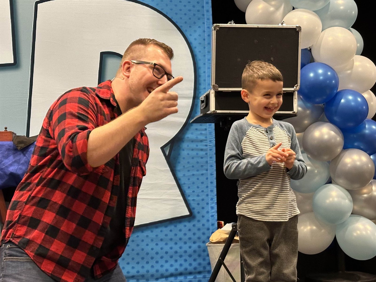 The birthday boy beams on stage under the balloon arch while Tyler points him out to the crowd