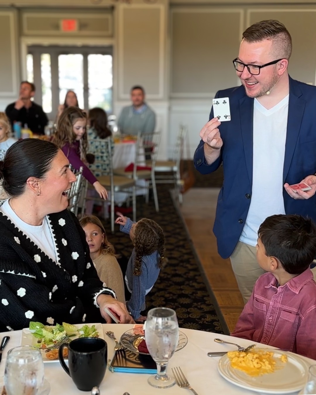 Tyler holds up a chosen card tableside as a club member laughs in disbelief over brunch