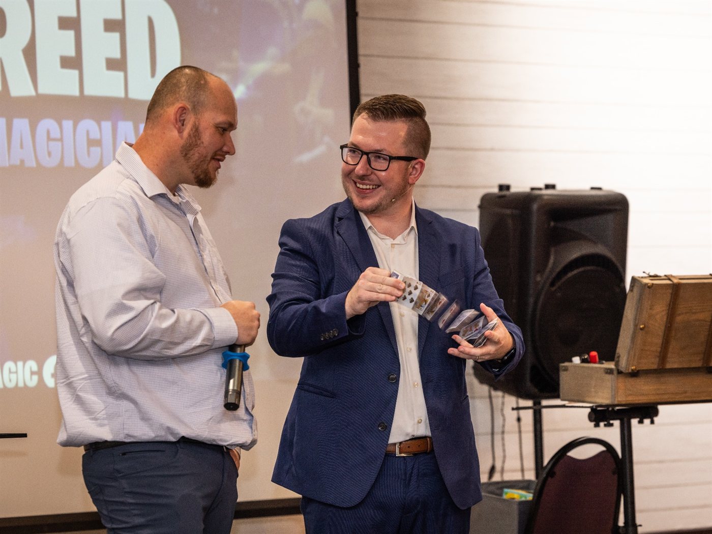 Tyler runs a card routine on stage beside a laughing volunteer at a company party