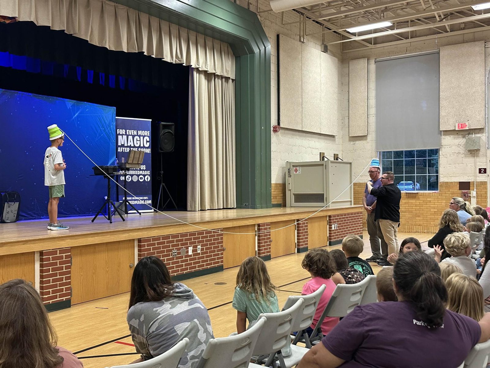 A packed community room watches a kid balance a bucket on his head on stage during the family show