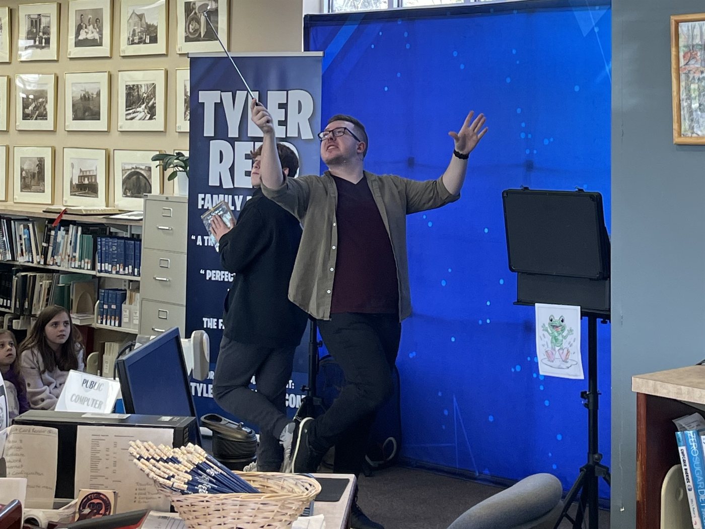 A young volunteer raises the magic wand beside Tyler in the middle of the library stacks
